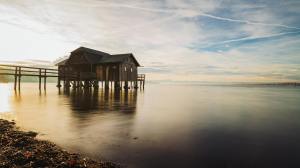 A serene lakeside scene featuring a wooden structure on stilts, surrounded by calm water and a picturesque sky with soft clouds.