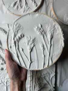 A hand holding a round, textured piece of white clay or plaster with floral designs, depicting tall flowers or plants against a smooth backdrop.