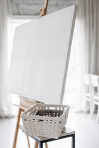 A blank canvas on an easel with a white basket underneath it.
